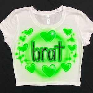 May include: White crop top with a green airbrushed design. The design features the word "brat" in black letters surrounded by green hearts and stars.