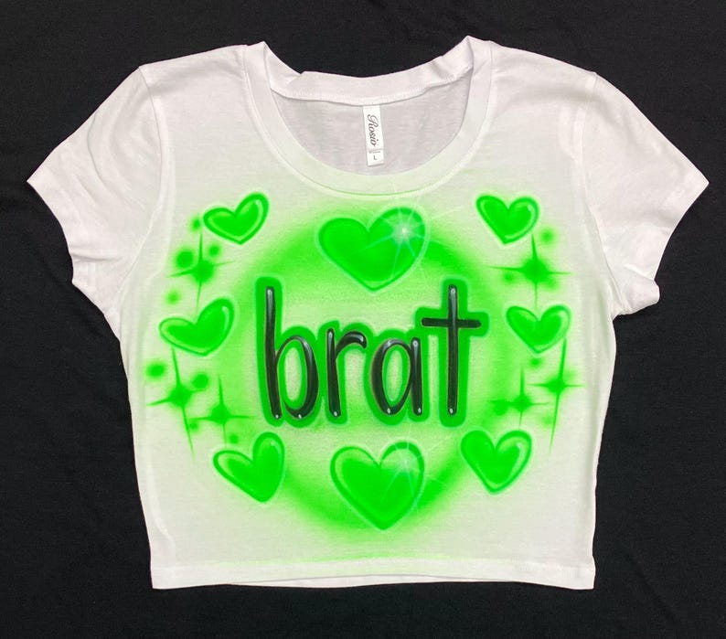 May include: White crop top with a green airbrushed design. The design features the word "brat" in black letters surrounded by green hearts and stars.
