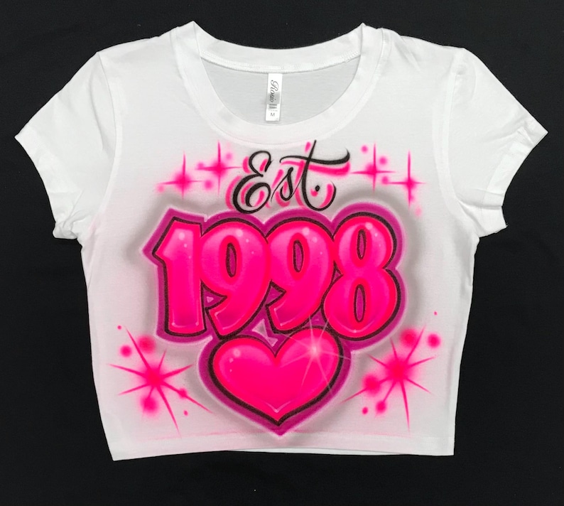 May include: White crop top with a pink airbrushed design. The design features the year 1998 in large, bubble letters and the word "Est" above it. A pink heart is below the year.