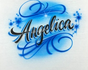 May include: A white t-shirt featuring an airbrushed design with the name Angelica written in black cursive script. The text is surrounded by blue swirling flourishes, soft blue shading, and several small blue star-shaped accents.