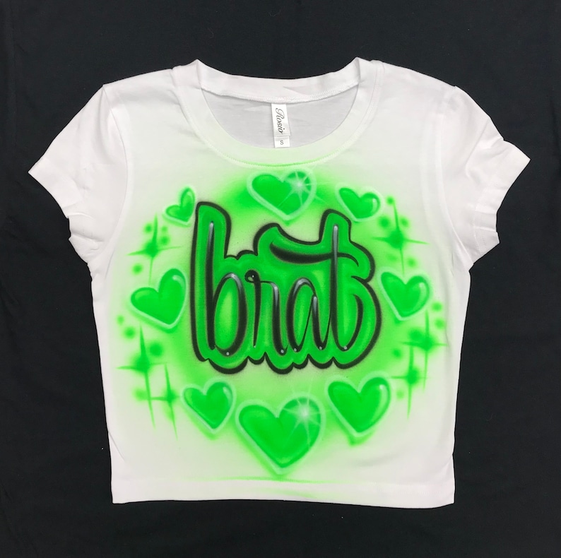 May include: White crop top with a green airbrushed design. The design features the word "brat" in a stylized font surrounded by green hearts.