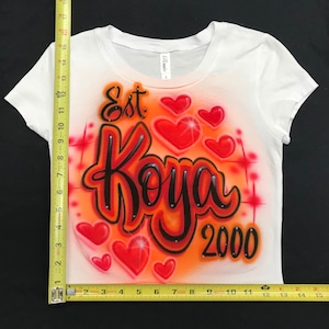 May include: White t-shirt with an orange and red airbrushed design. The design features the text "Est Koya 2000" with several red hearts.