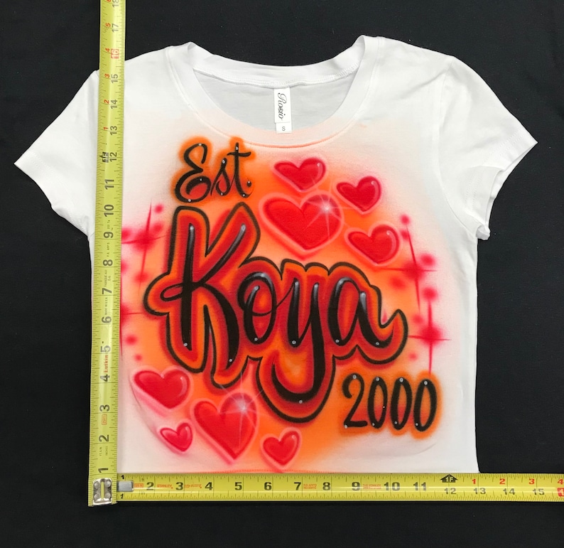 May include: White t-shirt with an orange and red airbrushed design. The design features the text "Est Koya 2000" with several red hearts.