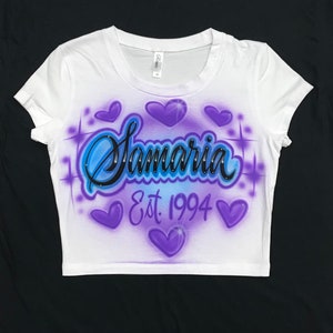 May include: White crop top with a purple and blue airbrushed design. The design features the name "Samaria" with rhinestones, a heart, and the text "Est. 1994".