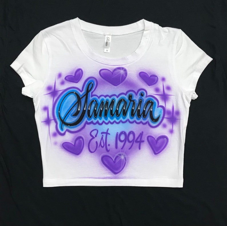 May include: White crop top with a purple and blue airbrushed design. The design features the name "Samaria" with rhinestones, a heart, and the text "Est. 1994".