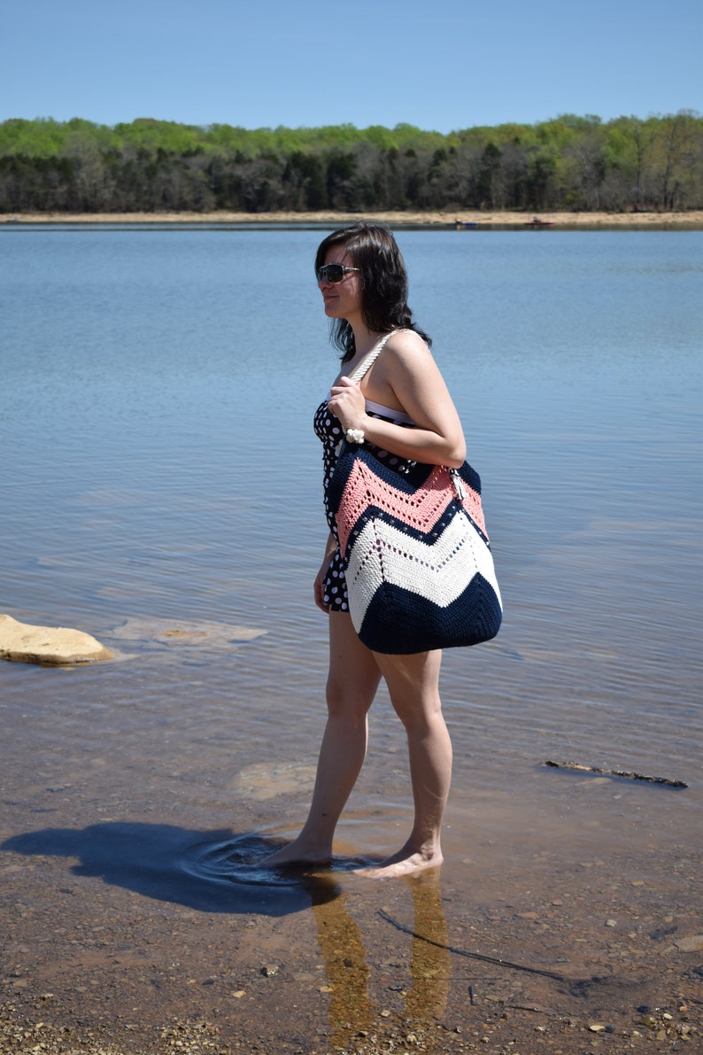 K&ouml;nnte beinhalten: Eine Frau in einem gepunkteten Badeanzug und Sonnenbrille steht im seichten Wasser und h&auml;lt eine gro&szlig;e geh&auml;kelte Strandtasche mit rosa, wei&szlig;en und dunkelblauen Streifen.
