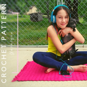 Puede incluir: Una persona joven con leggings azules y un top amarillo sin mangas se sienta en una alfombra rosa de crochet con un perro negro pequeño. Lleva auriculares azules y escucha música.