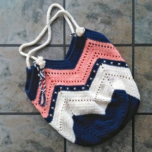 K&ouml;nnte beinhalten: Eine geh&auml;kelte Tragetasche mit einem Chevron-Muster in Rosa, Wei&szlig; und Marineblau. Die Tasche hat einen wei&szlig;en Quasten und silberne Nieten.