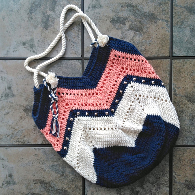 K&ouml;nnte beinhalten: Eine geh&auml;kelte Tragetasche mit einem Chevron-Muster in Rosa, Wei&szlig; und Marineblau. Die Tasche hat einen wei&szlig;en Quasten und silberne Nieten.