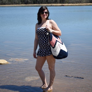 K&ouml;nnte beinhalten: Eine Frau in einem schwarz-wei&szlig;en Polka-Dot-Badeanzug steht im seichten Wasser und h&auml;lt eine rosafarbene, wei&szlig;e und blaue geh&auml;kelte Strandtasche.