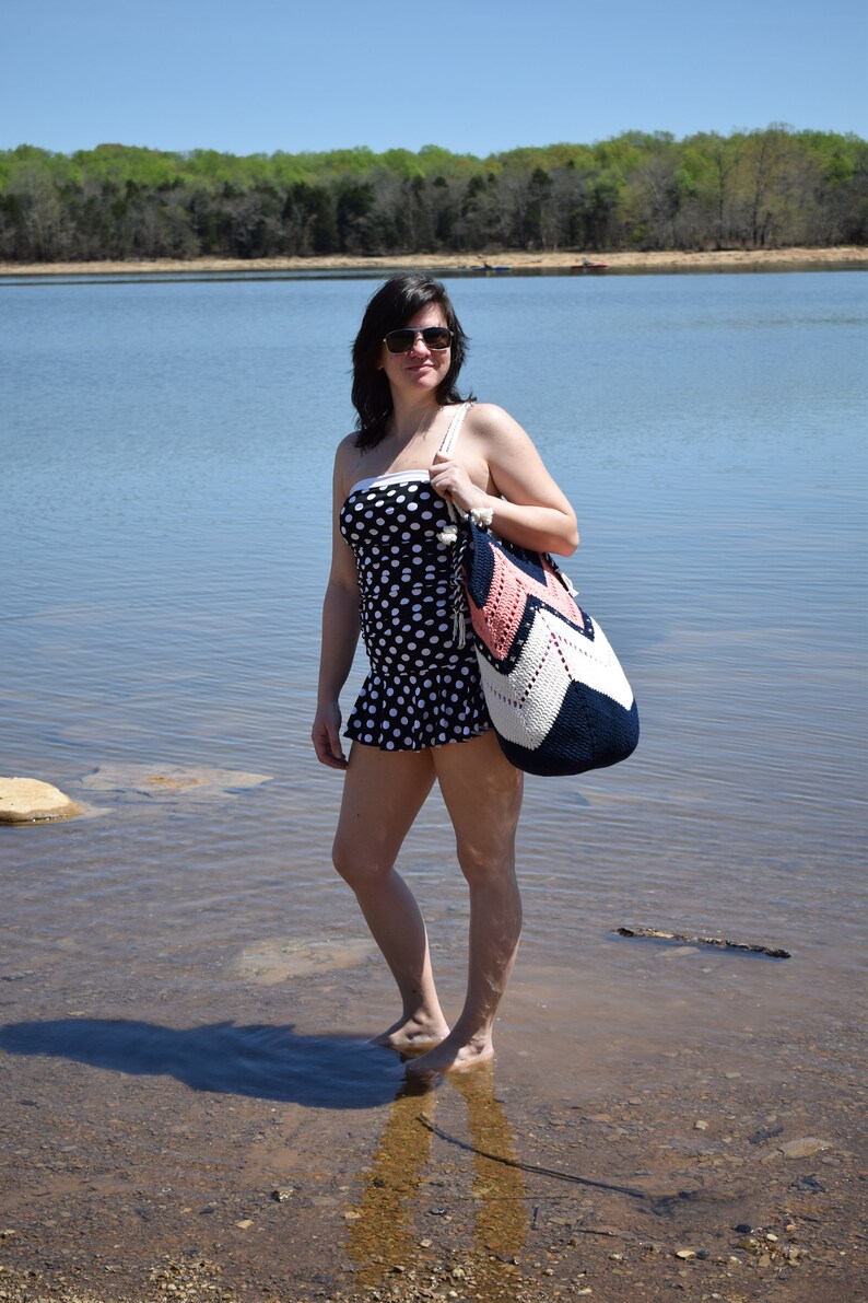 K&ouml;nnte beinhalten: Eine Frau in einem schwarz-wei&szlig;en Polka-Dot-Badeanzug steht im seichten Wasser und h&auml;lt eine rosafarbene, wei&szlig;e und blaue geh&auml;kelte Strandtasche.