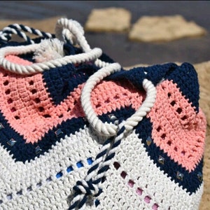 K&ouml;nnte beinhalten: Eine geh&auml;kelte Tragetasche mit einem geometrischen Muster in Wei&szlig;, Rosa und Marineblau. Die Tasche hat einen wei&szlig;en Kordelgriff und einen Kordelzug in Marineblau und Wei&szlig;.