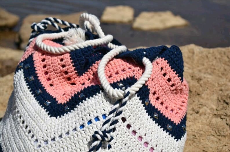 K&ouml;nnte beinhalten: Eine geh&auml;kelte Tragetasche mit einem geometrischen Muster in Wei&szlig;, Rosa und Marineblau. Die Tasche hat einen wei&szlig;en Kordelgriff und einen Kordelzug in Marineblau und Wei&szlig;.