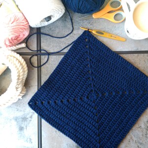 K&ouml;nnte beinhalten: Ein quadratisches H&auml;kelst&uuml;ck in Marineblau mit einem wei&szlig;en Garnrand. Das Quadrat ist mit einem einfachen Stichmuster gearbeitet und hat eine strukturierte Optik.