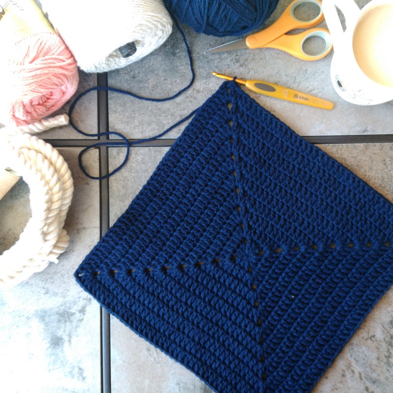 K&ouml;nnte beinhalten: Ein quadratisches H&auml;kelst&uuml;ck in Marineblau mit einem wei&szlig;en Garnrand. Das Quadrat ist mit einem einfachen Stichmuster gearbeitet und hat eine strukturierte Optik.
