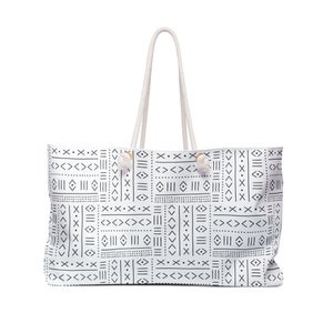 May include: White tote bag with a black geometric pattern. The bag has a rectangular shape with a cream-colored rope handle. The design features lines, dots, and arrow-like shapes. The bag is designed for carrying items.