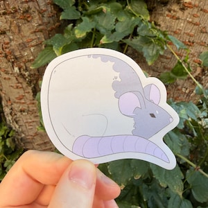 May include: A die-cut sticker featuring a stylized illustration of a curled-up mouse. The mouse has a white body, a light purple tail, and a grey head with light purple inner ears. The design is outlined in black with a white border.