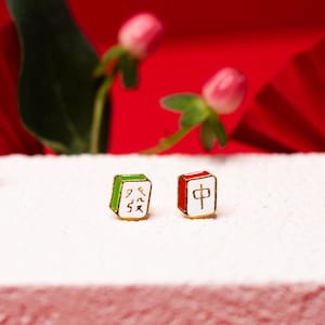 May include: A pair of gold-tone earrings shaped like mahjong tiles. The left earring is green and white with Chinese characters, and the right earring is red and white with the Chinese character for "middle".