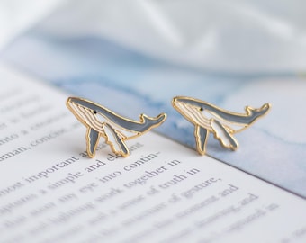May include: A pair of gold-toned metal stud earrings shaped like humpback whales. Each whale features a smooth grey enamel body with white underbelly detailing and a gold outline, capturing the distinct silhouette of the marine mammal.