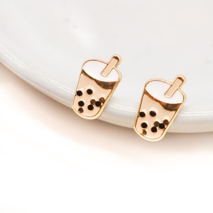 May include: A pair of gold-tone stud earrings featuring a white enamel boba tea cup with black boba pearls and a gold straw.
