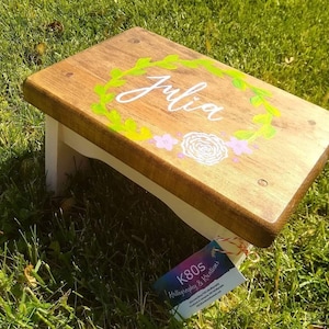 May include: A wooden step stool with a light brown top and white legs. The top is decorated with the name "Julia" in white script, surrounded by a green wreath and pink and white flowers. The stool is on a grassy surface.