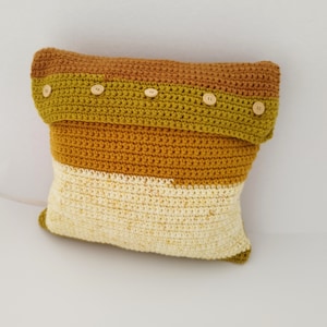 Könnte beinhalten: Ein gehäkeltes Kissen mit einem gestreiften Design in Senfgelb, Olivgrün und Braun. Das Kissen hat eine Klappe mit Holzknöpfen.