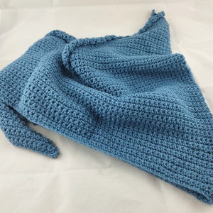 May include: A blue crochet shawl, folded and draped. The shawl is a solid color, with a textured stitch pattern and a scalloped edge. The handmade shawl is made of soft yarn.