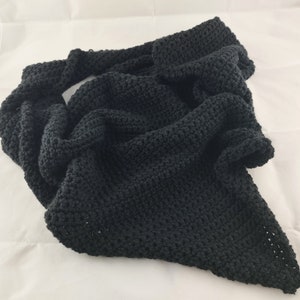 May include: A black knitted scarf, draped on a white surface. The scarf is made of a thick, textured yarn, and is folded to create a layered effect. The scarf's dark color contrasts with the white background.