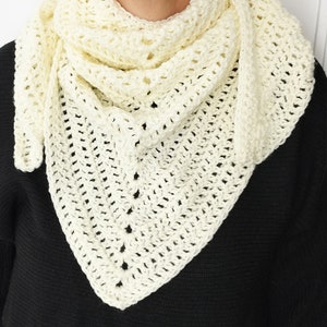 May include: Cream-colored, handmade crochet shawl. The triangular shawl is draped around the neck and shoulders. The open weave design features a series of small holes throughout the fabric. The shawl is made of a soft, textured yarn.