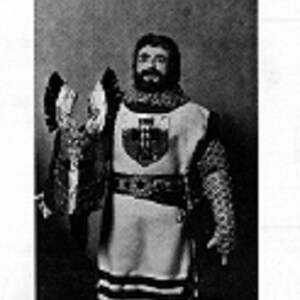 May include: A black and white image of a person in medieval armor. The armor includes a chainmail coif, sleeves, and leggings. The person wears a tunic with a shield emblem and holds a shield.