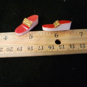 Könnte beinhalten: Zwei Miniatur-Sandalen in Rot und Gold mit einem goldenen Band oben. Die Sandalen stehen auf einer weißen und rosa Basis. Ein Holzlineal befindet sich im Hintergrund.