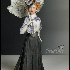 Könnte beinhalten: Puppe in einem Outfit im Vintage-Stil, die einen weißen Spitzen-Sonnenschirm hält. Die Puppe trägt einen grauen Rock, eine weiße Bluse und ein kariertes Cape. Ein Hut mit floralen Akzenten vervollständigt das Ensemble. Die Puppe hat einen ernsten Gesichtsausdruck.