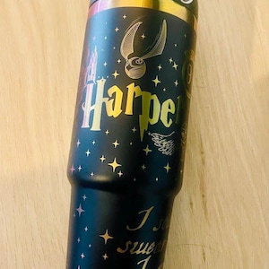 Könnte beinhalten: Schwarzer isolierter Becher mit goldenen Akzenten und einem Griff. Der Becher hat goldfarbene Schriftzüge mit der Aufschrift "Harper" und ein goldenes Schnatz-Design. Der Becher hat auch den Text "I solemnly swear I am up to no good."