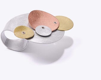 Pode incluir: Um anel com uma banda larga de prata escovada rematada por um disco grande e plano de prata. Sobre esta base, encontram-se três elementos circulares mais pequenos e sobrepostos em ouro, cobre e prata escovados, cada um fixado com um pequeno rebite.