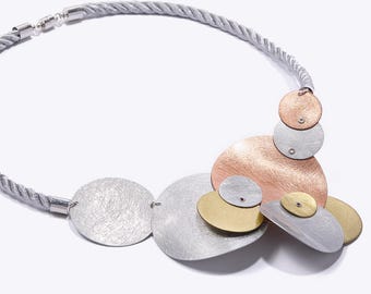 Pode incluir: Um colar com um cordão torcido em tom prateado e um grupo de discos metálicos circulares sobrepostos em tons de prata escovada, cobre e ouro. Os discos estão ligados por pequenos rebites metálicos, criando um design de pendente assimétrico.