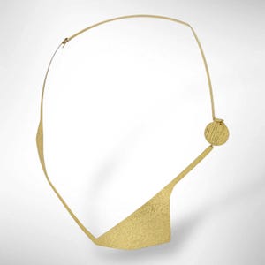 May include: A gold-colored necklace with a unique, abstract design. The necklace features a curved upper portion, a textured triangular pendant, and a circular accent. The metal has a hammered texture, giving it a handcrafted appearance.