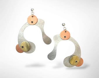 Pode incluir: Um par de brincos abstratos e modernos. Cada brinco apresenta uma forma metálica curva prateada, um pequeno disco redondo cor de cobre no topo e um disco dourado e um disco cor de cobre na parte inferior. Penduram de uma pequena bola prateada.