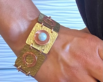 Pode incluir: Uma pulseira artesanal com elos quadrados de metal texturizado em tons de ouro, cobre e prata. Cada elo quadrado é conectado por pequenos anéis de metal. O elo central tem um círculo de cobre com uma pedra azul clara.