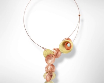 Puede incluir: Un collar único con una fina cadena de alambre de cobre. El colgante presenta una serie de discos y esferas de metal mixto en tonos dorados y cobrizos. Una pequeña piedra azul claro está engastada en el disco más grande. Diseño moderno y artístico.