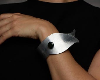 Pode incluir: Uma pulseira larga de metal prateado escovado com uma silhueta curva em forma de folha que envolve o pulso. O design é acentuado com uma única pedra preta pequena, lisa e redonda, centralizada na superfície metálica.