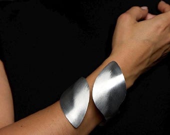 Pode incluir: Uma pulseira de punho prateada com acabamento em metal escovado. A pulseira apresenta um design dividido com dois grandes elementos em forma de folha. A joia é usada no pulso de uma pessoa, contra um fundo preto.
