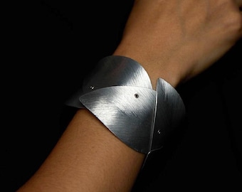 Pode incluir: Uma pulseira larga tipo bracelete feita de placas de metal prateado em forma de pétalas sobrepostas, com um acabamento escovado e fosco. Os segmentos individuais são fixados com pequenos rebites visíveis, criando uma estrutura orgânica em camadas.