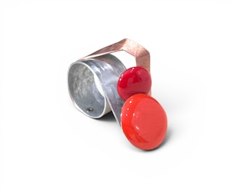 Puede incluir: Un anillo con una banda ancha de tono plateado texturizado que se curva hacia arriba, rematado con un elemento angular de color cobre. Dos acentos circulares lisos y brillantes están unidos al frente, uno en rojo oscuro y otro en naranja rojizo.