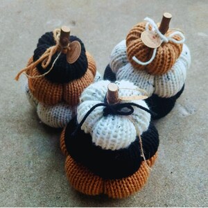 May include: Three stacked knit pumpkins in shades of black, orange, and white, topped with wooden stems. The text "STACKED PUMPKINS" is above the pumpkins, and "LULA BILL BOUTIQUE" is below.
