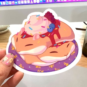 May include: A whimsical sticker featuring a stack of cartoon hamsters designed to look like a dessert. The hamsters are light orange with pink ears, topped with pink frosting, sprinkles, berries, and a drizzle of red sauce, all on a purple plate.