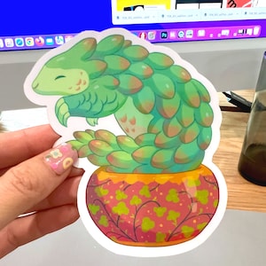 May include: A sticker featuring a green pangolin-like creature with a smiling face, nestled in a decorative pot. The pangolin has overlapping scales in shades of green and orange. The pot is red with a floral pattern.