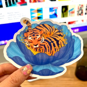 Puede incluir: Una pegatina con un tigre durmiente con rayas naranjas y negras acurrucado en una flor azul. La flor tiene detalles en azul claro y blanco. El tigre está acurrucado con los ojos cerrados, dando un ambiente de paz.