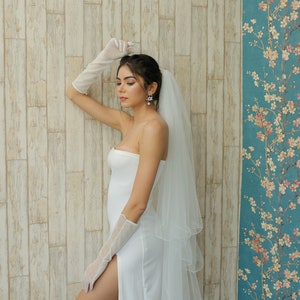 Puede incluir: Una mujer con un vestido de novia blanco, velo y guantes blancos. Está de pie frente a una pared de madera con paneles y un papel tapiz floral detrás de ella.