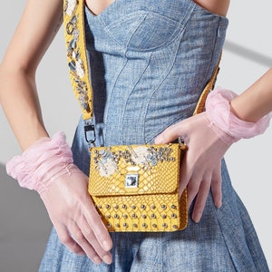 May include: A yellow crossbody bag with a snakeskin pattern and silver embellishments. The bag has a matching yellow strap with decorative elements. The person is wearing pink gloves and a blue denim dress.