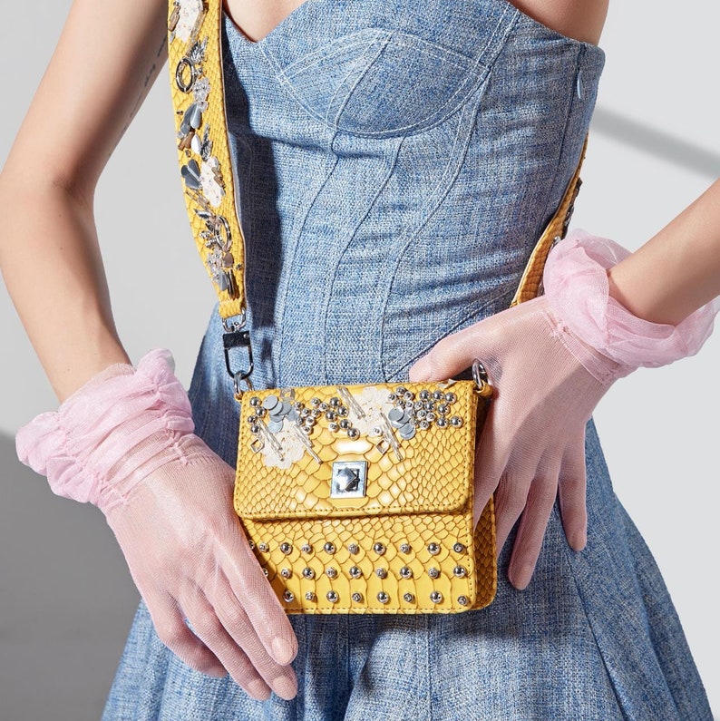 May include: A yellow crossbody bag with a snakeskin pattern and silver embellishments. The bag has a matching yellow strap with decorative elements. The person is wearing pink gloves and a blue denim dress.